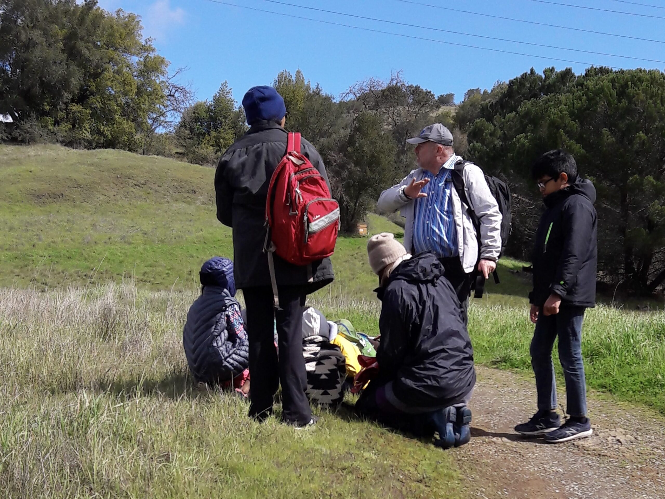 Instructor, parents, and kids discussing and learning about the biogeomorphology at a park. Many of the young people and one parent are kneeling or sitting on the ground listening and observing.