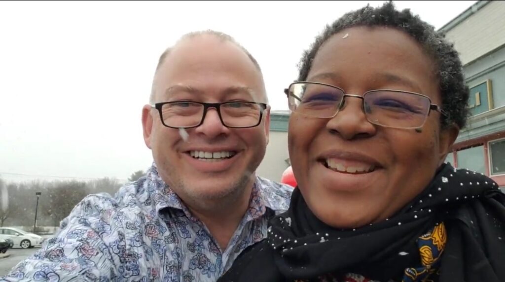 A Black woman with short natural hair and partially bald white man laugh as they take a picture as snow falls around them. He is wearing a white, red, and blue floral button-up shirt with the top buttons open and they are wearing a dark floral dress wit a sparkling scarf around their neck. Both of them wear glasses, sport laugh creases, and apple cheeks from their big smiles as they laugh.